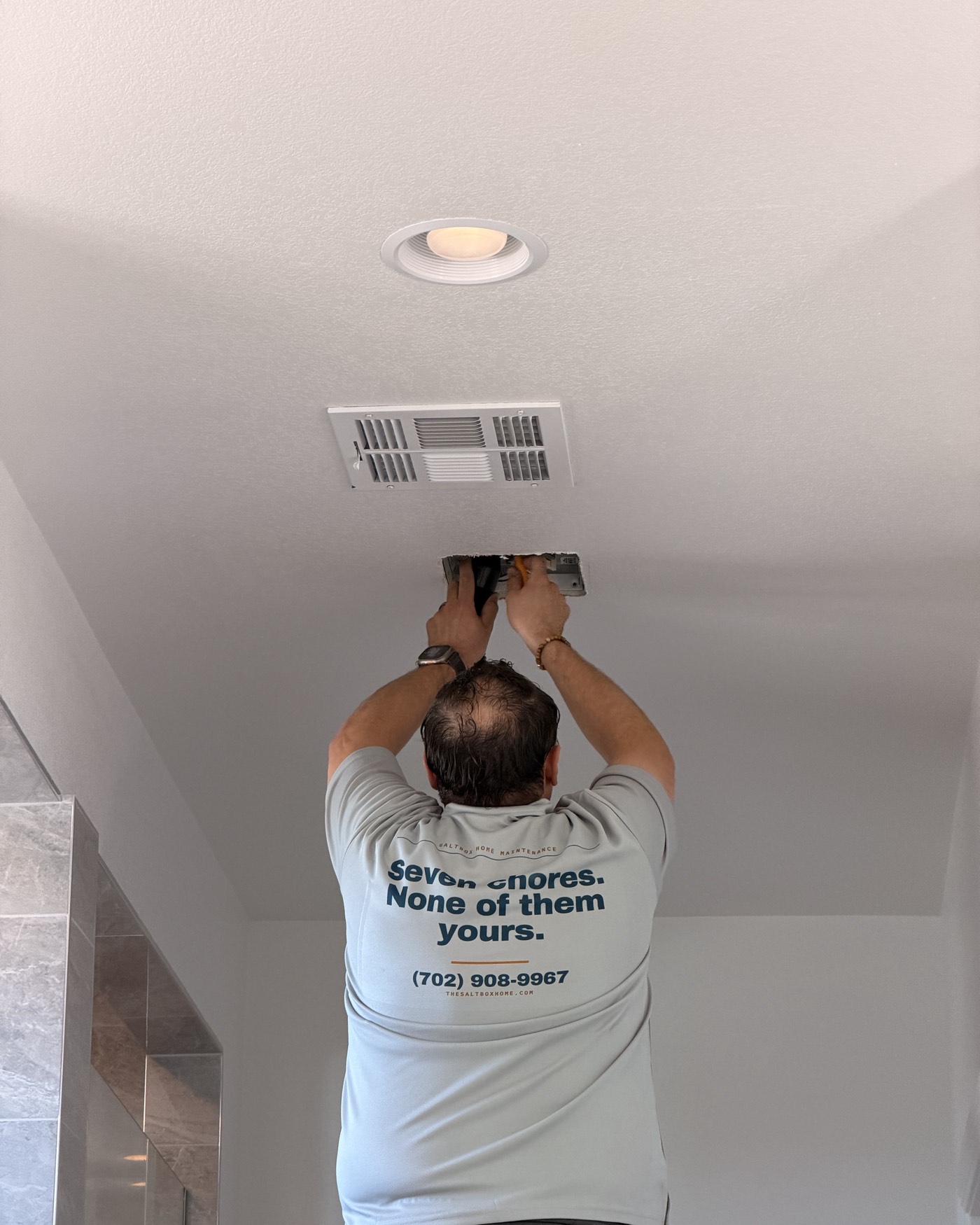 A technician in a Saltbox polo reaching up to release the grille of a ceiling bathroom exhaust fan